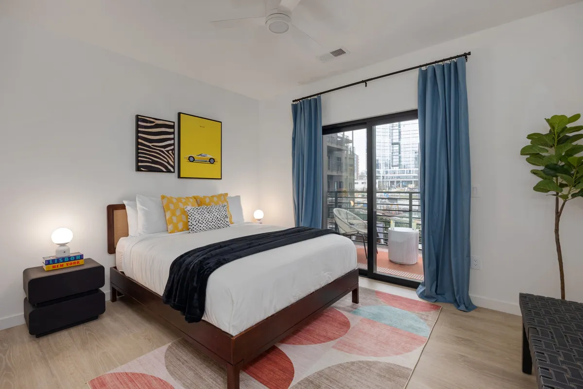 A stylish Queen bedroom with decorative pillows, a large window that opens to a balcony overlooking the pool area, vibrant artwork on the wall, and a cozy, colorful rug. The room exudes comfort and offers easy outdoor access.