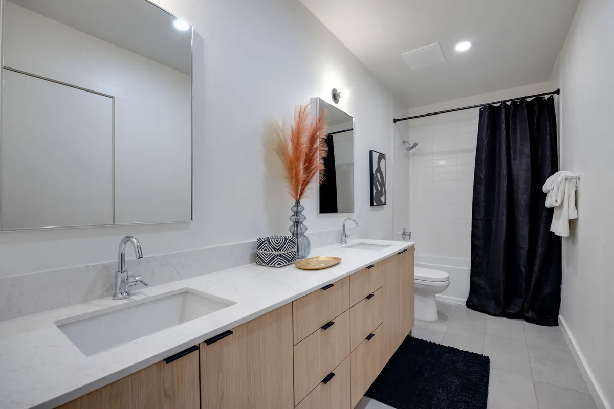 A serene designer bathroom featuring dual sinks, large mirrors, decorative pampas grass, and a sleek black shower curtain. The space feels spa-inspired, ideal for morning routines or relaxing self-care rituals.