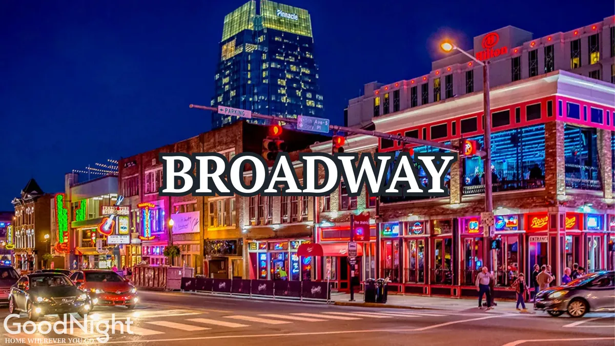 A bustling Nashville street at night, drenched in the glow of neon signs and lined with lively crowds, entertainment venues, cars, and shops. The energy is high and the atmosphere is unmistakably urban and vibrant, perfectly capturing the nightlife of Broadway.