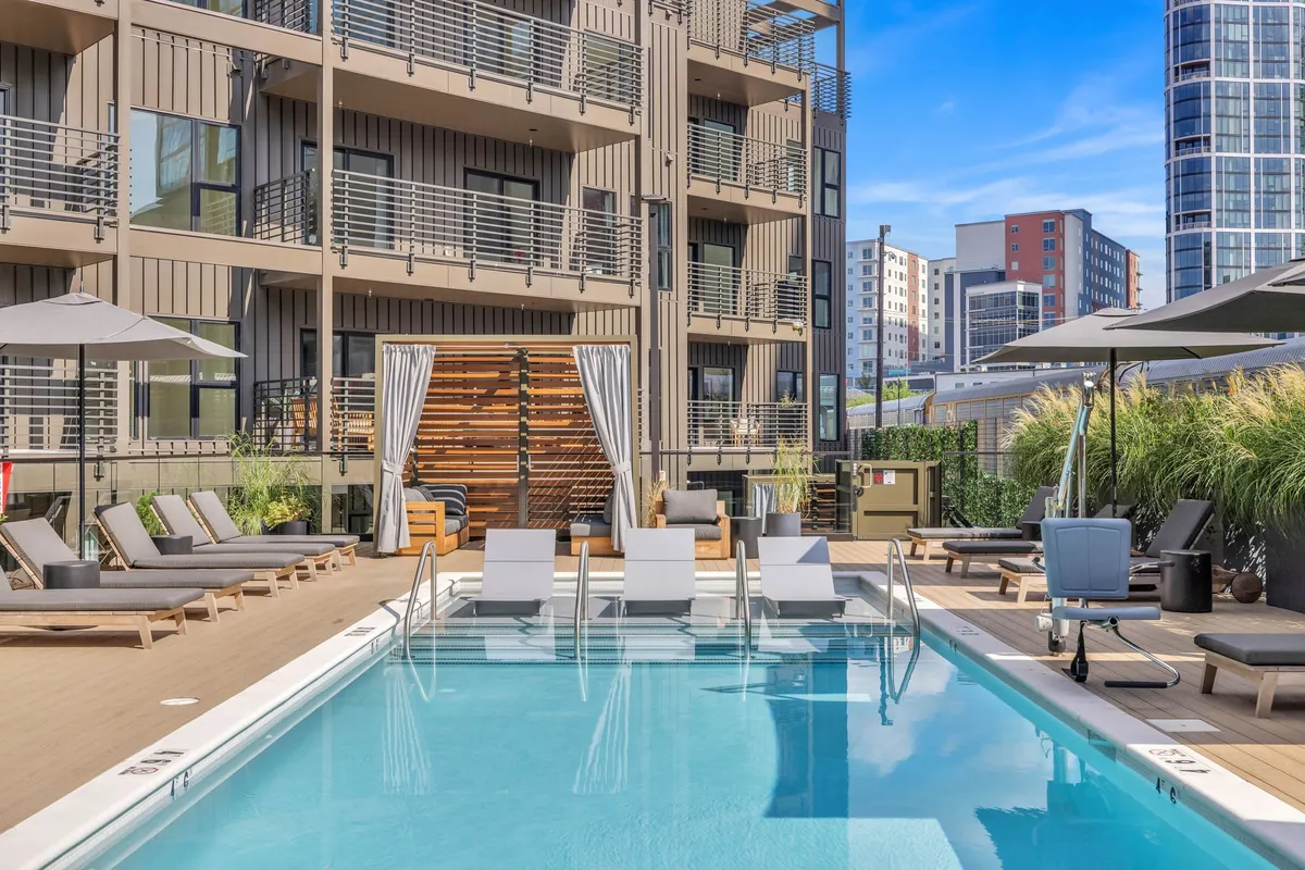 An inviting outdoor swimming pool area with lounge chairs and sun umbrellas in front of a modern building facade. The setting is social and upscale, hinting at resort-style amenities.