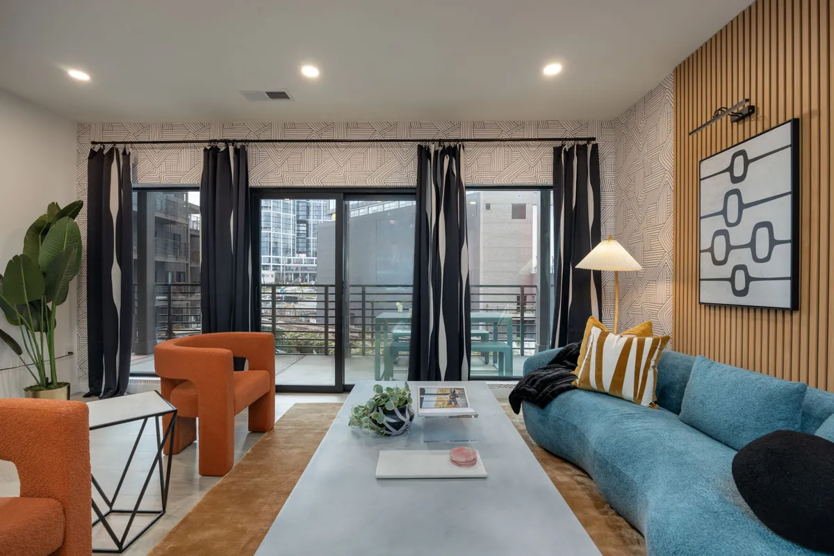 The living area features a blue sectional sofa, bright orange accent chairs, a modern geometric coffee table, and large windows with city views. The abundance of sunlight and bold color choices evoke a bohemian-chic vibe, making the space feel lively and perfect for both relaxation and social gatherings.