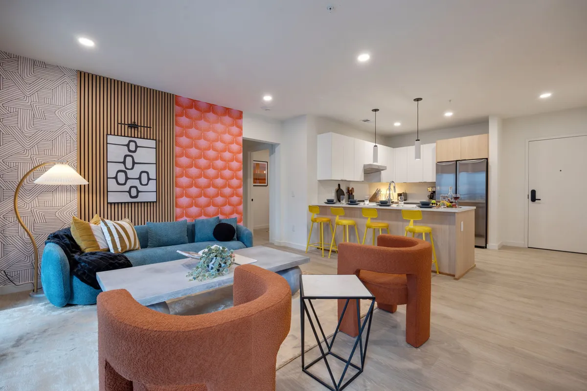 The image showcases a vibrant living space with a colorful accent wall, a sleek modern kitchen island, and a cozy seating area illuminated by a decorative lamp. The atmosphere is lively and welcoming, perfect for socializing, cooking, and relaxing with friends or family.
