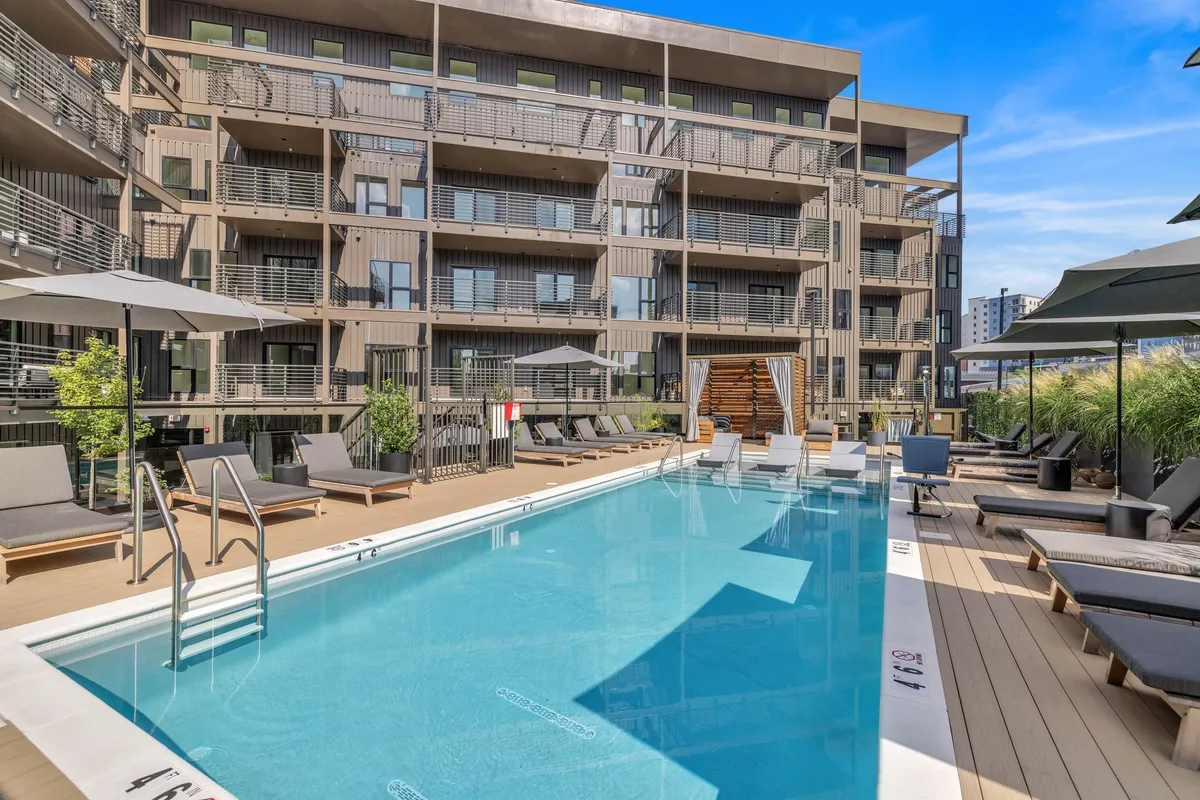 This image features an inviting swimming pool surrounded by lounge chairs, with the modern building facade and lush greenery in the background. The layout suggests a resort-style area ideal for relaxation and group gatherings.