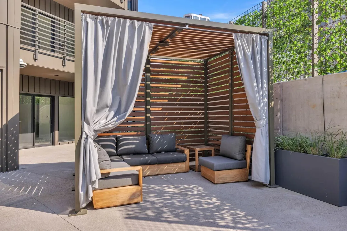 A tranquil rooftop lounge space with a wooden pergola, comfortable seating, soft curtains, and lush greenery in planters. The area is designed for relaxing, socializing, and enjoying the city views in a cozy, upscale setting.