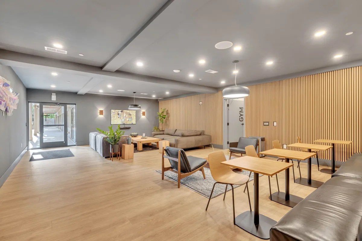 A vibrant interior lounge area featuring comfortable sofas, dining tables, wall art, potted plants, and a gym door in the background. The modern decor and communal layout evoke a boutique hotel atmosphere, inviting guests to gather and socialize.