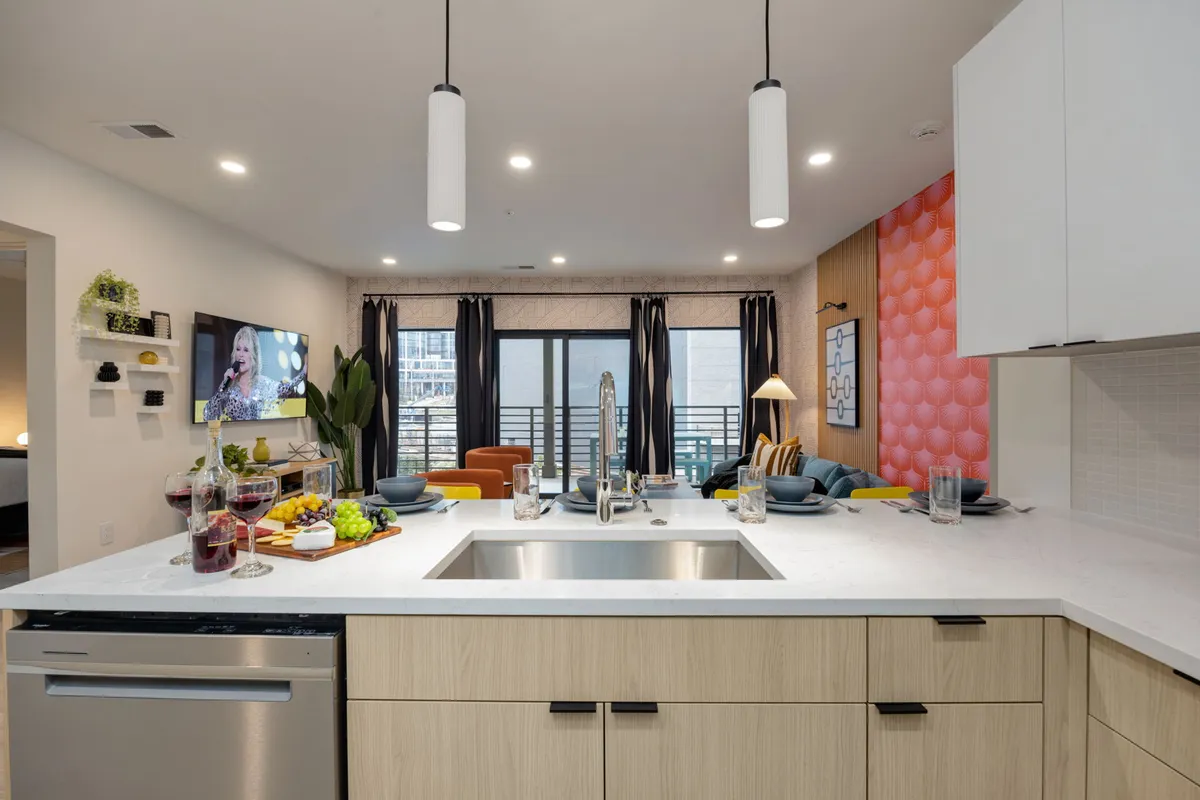 A wide-angle view captures the open-concept living, dining, and kitchen area. The kitchen island anchors the space, flanked by a dining setup and a cozy living room arrangement bathed in natural light from large windows. Designer furnishings and vibrant colors infuse the room with energy and a welcoming ambiance ideal for gatherings.