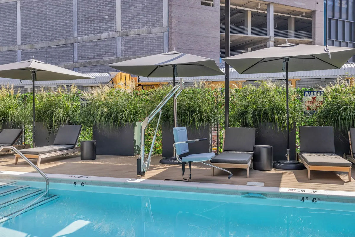 A spacious resort-style swimming pool surrounded by inviting lounge chairs, umbrellas, and tall grass planters. The pool area is bathed in natural light, and the mood is calm and leisurely—guests can be seen sunbathing, swimming, and relaxing with drinks. The setup hints at opportunities for socializing and quiet moments alike.