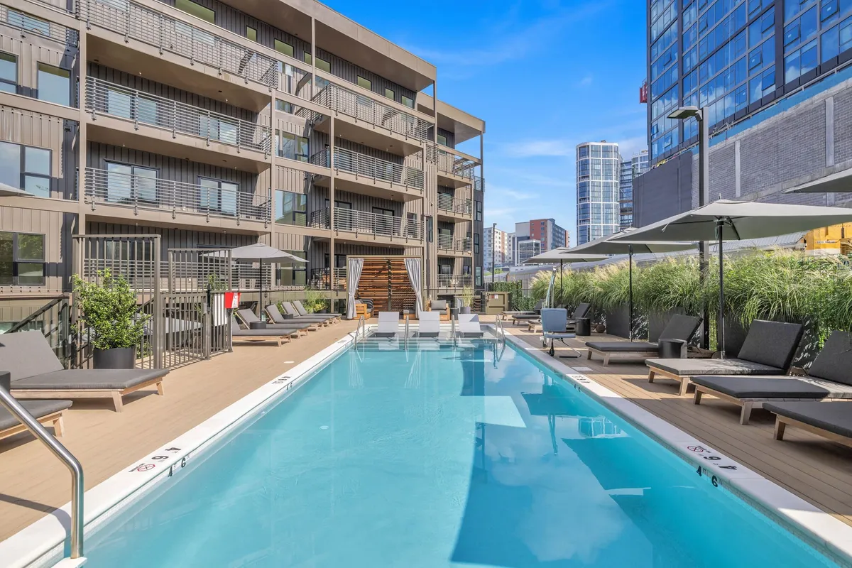 An inviting outdoor swimming pool surrounded by lounge chairs, with city buildings in the background. The mood is relaxed, ideal for sunbathing, swimming, socializing, or unwinding poolside in an urban oasis.