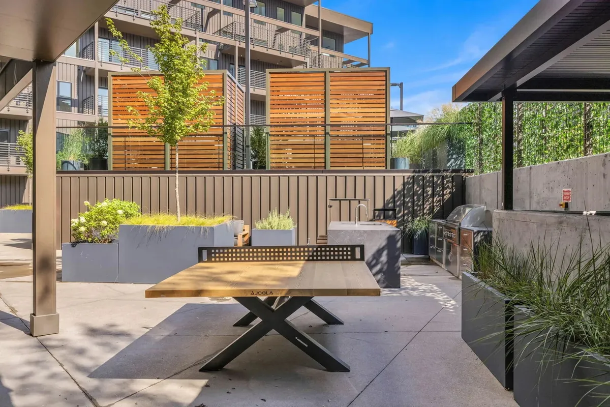 A stylish outdoor grilling and dining area, highlighted by a wooden table, built-in grill, decorative wooden screens, and abundant greenery. The space invites casual socializing, barbecuing, and outdoor dining against a backdrop of warm evening light.