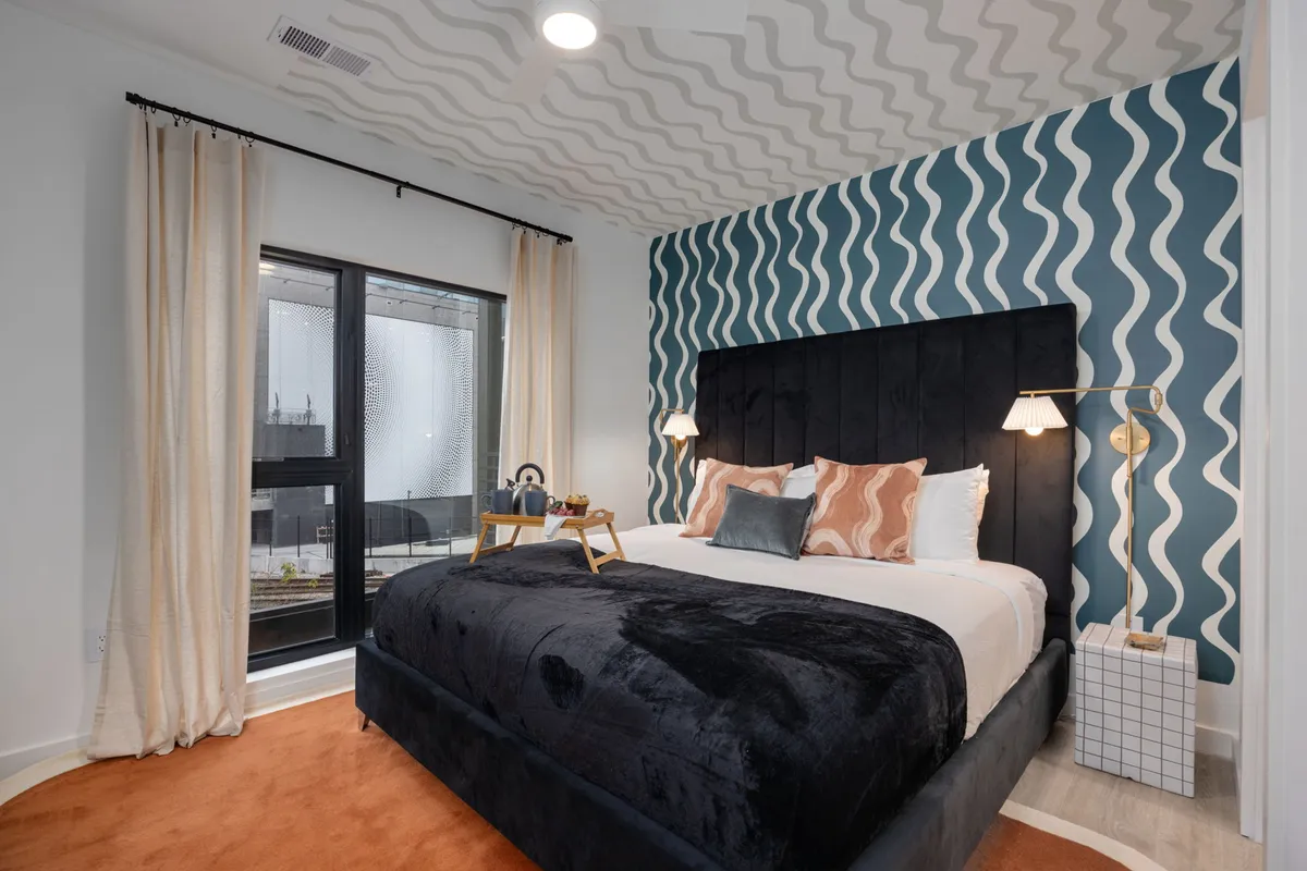 A modern bedroom featuring a comfortable bed against an accent wall, with a large window allowing city views and natural light to flood the space. The decor is inviting and contemporary, echoing a restful and rejuvenating atmosphere.